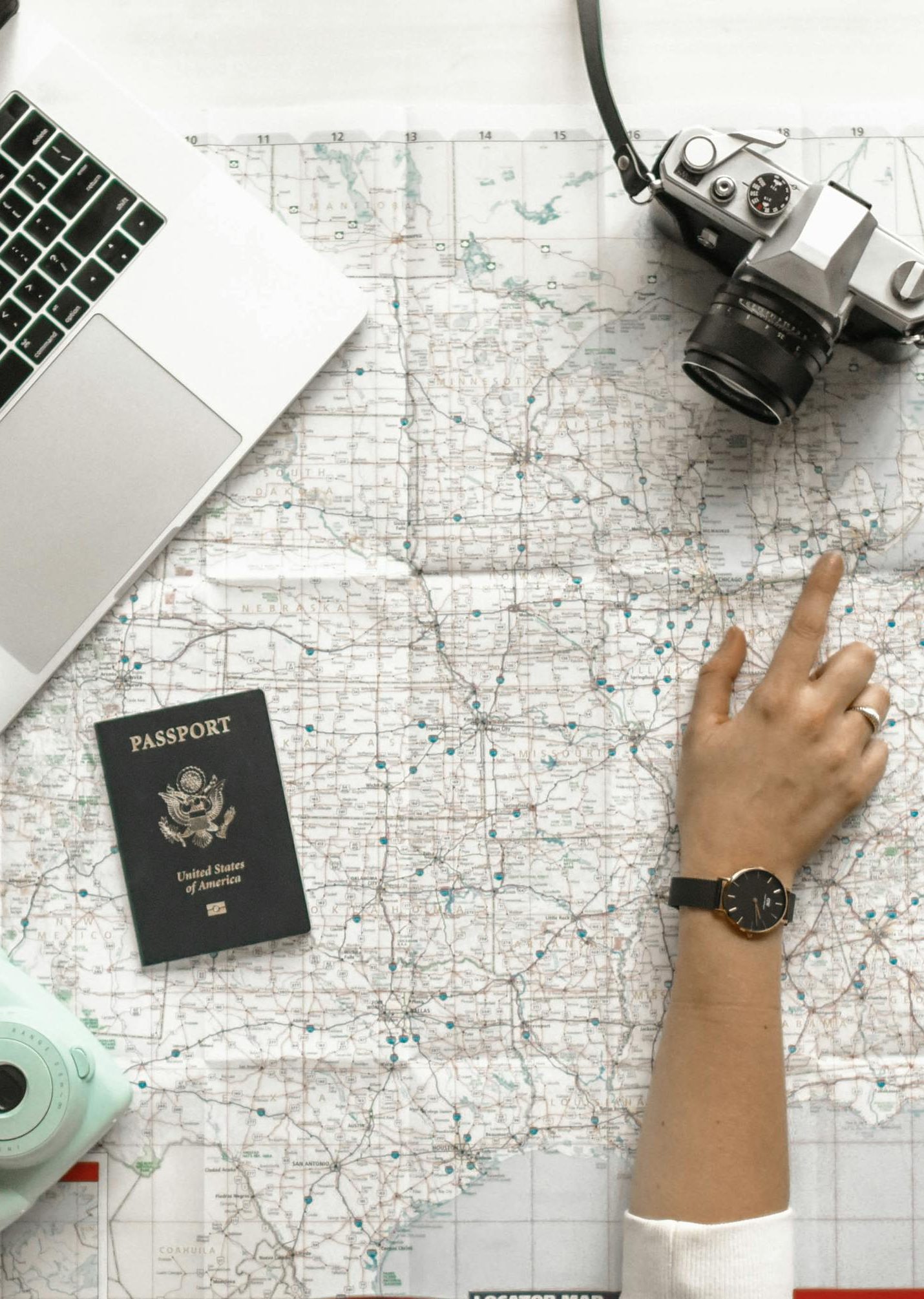 Hand on map with camera, passport, laptop, and coffee.